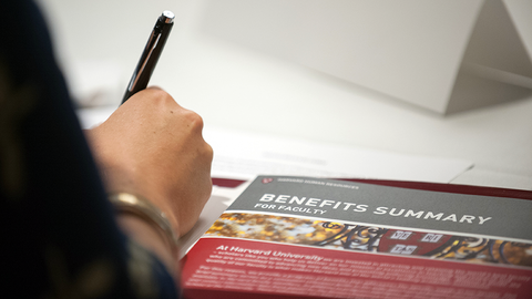 Close-up on a Faculty Benefits Summary Brochure with a hand holding a pen in the foreground.