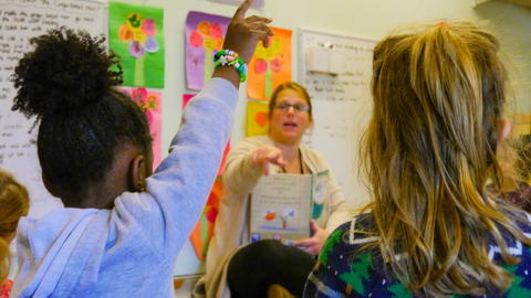 Two children in the foreground and a teacher in the background sitting on the floor in a classroom.