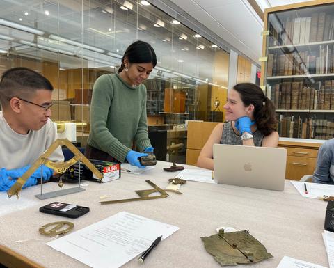 Students examining historical scientific instruments around a table.